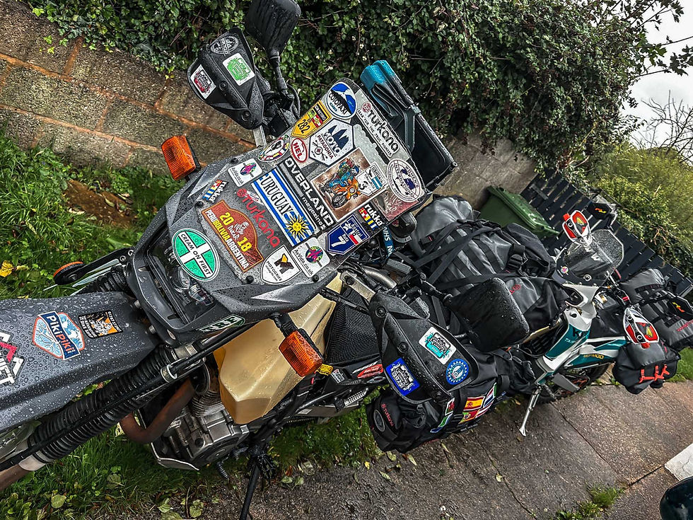 Motorcycles covered in travel stickers, parked by a hedge and stone wall. Turkana Gear Bags attached. Overcast and wet, evoking a feeling of adventure.