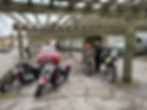Motorcycles and a red car are parked under a wooden pergola. A person adjusts gear on a bike. Street and buildings are visible in the background. AvVida micro adventures. Life is an adventure / La vida es Una Aventura!
