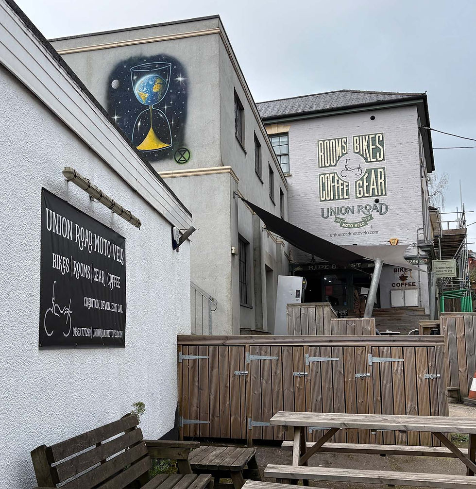 Outdoor view of Union Road Moto Velo. Signs advertise bikes, rooms, coffee, gear. Mural shows hourglass with Earth. Wooden benches/picnic tables. AvVida micro adventures. Life is an adventure / La vida es Una Aventura! Union Road Moto Velo