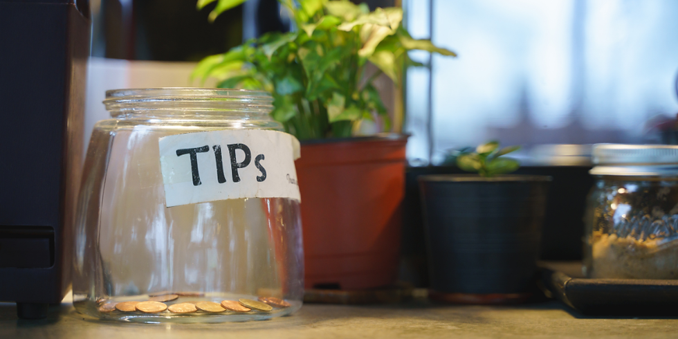 A glass tip jar filled with coins and notes sitting on a café counter, with a small sign inviting customers to leave tips.
