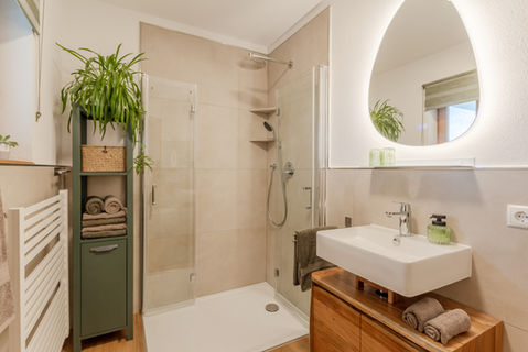 Neu gestaltetes Badezimmer in Beige und Grün mit großzügiger Dusche, Holzwaschtisch und beleuchtetem organischem Spiegel.