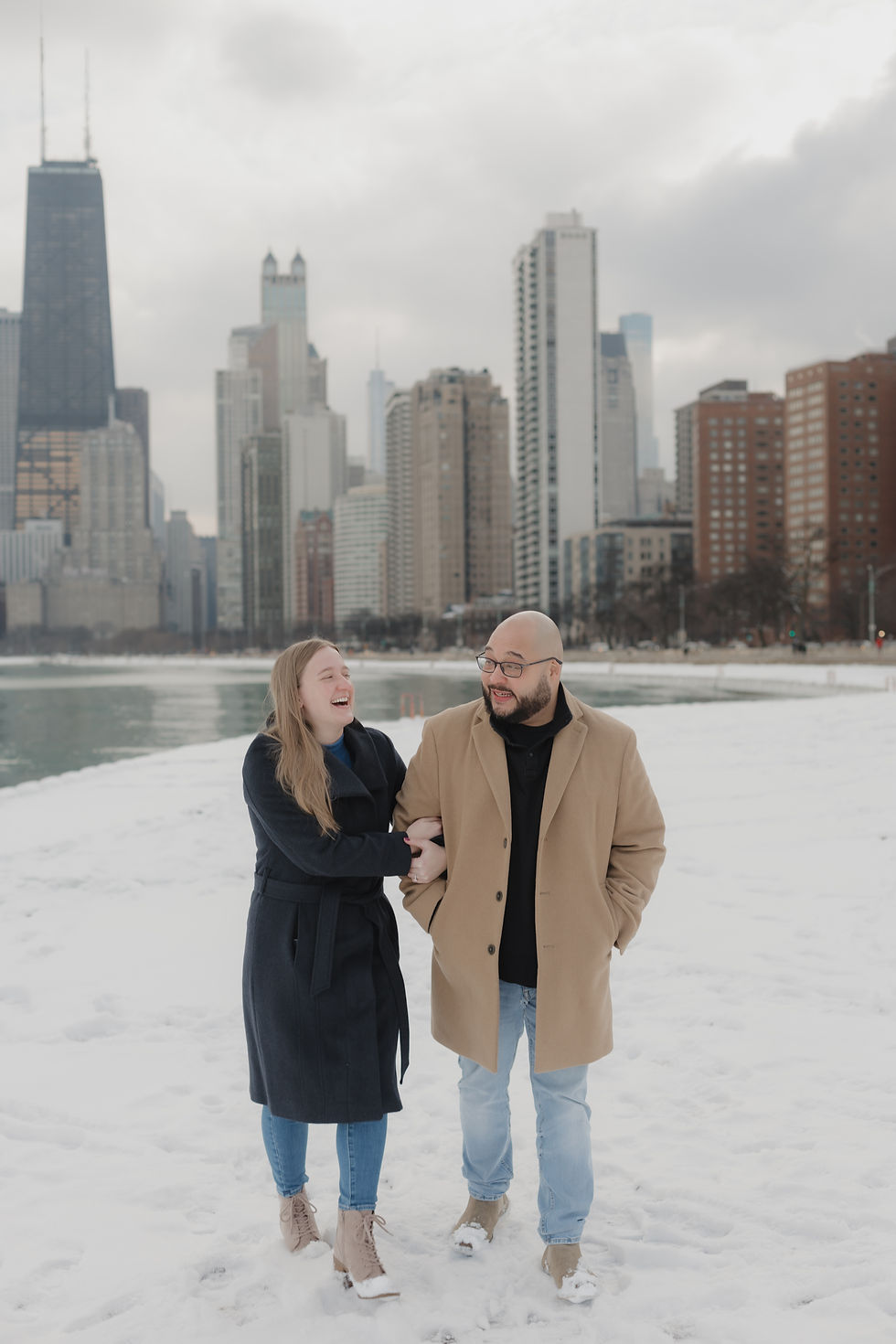 A couple laughs while walking on a snowy path with a city skyline in the background. The mood is joyful, and they wear winter coats.