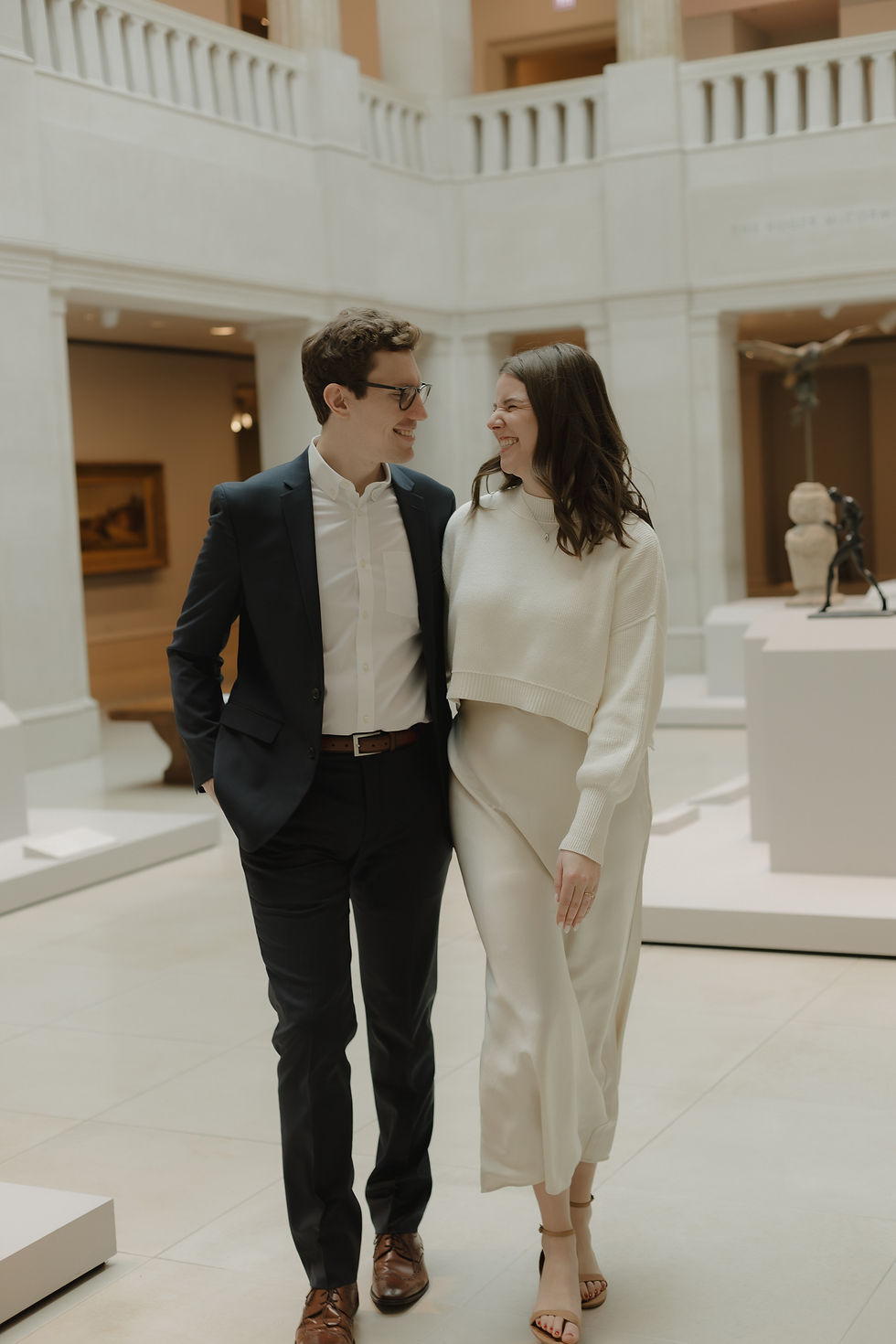 A couple walks arm-in-arm, smiling in a bright, modern gallery with white walls and sculptures.