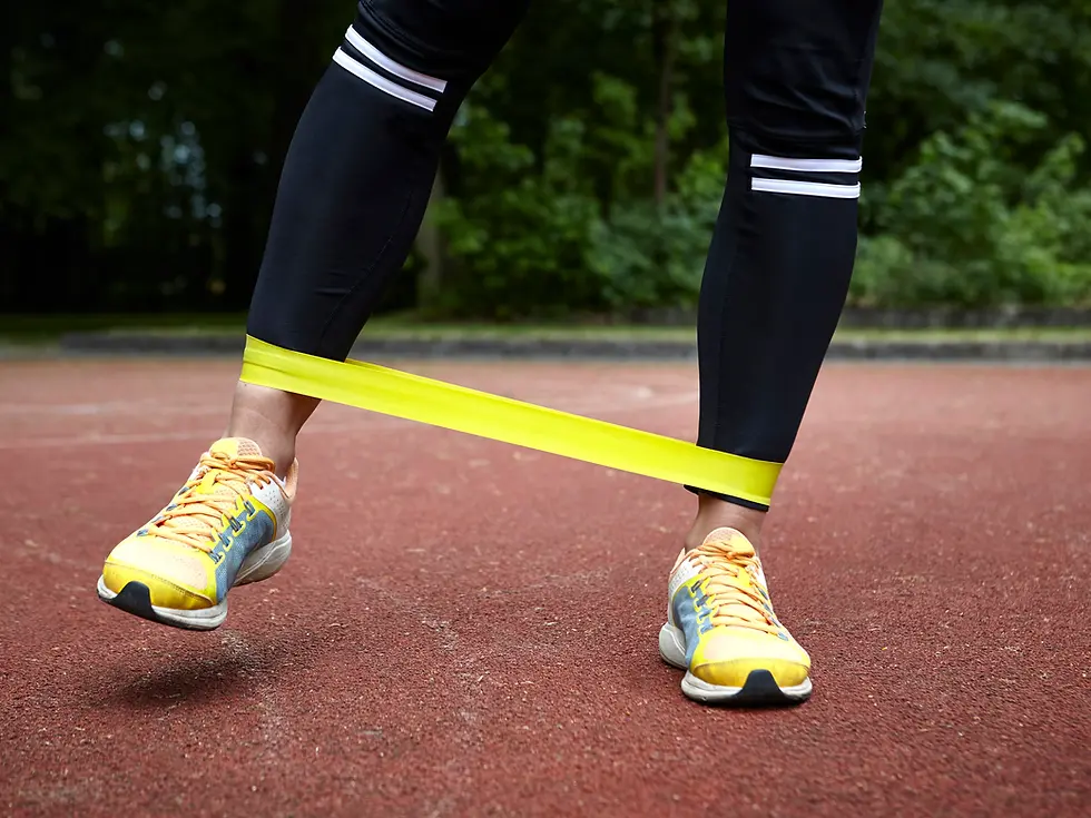 Person exercising with a yellow resistance band on a red track. Wearing black leggings and yellow sneakers. Green trees in the background.