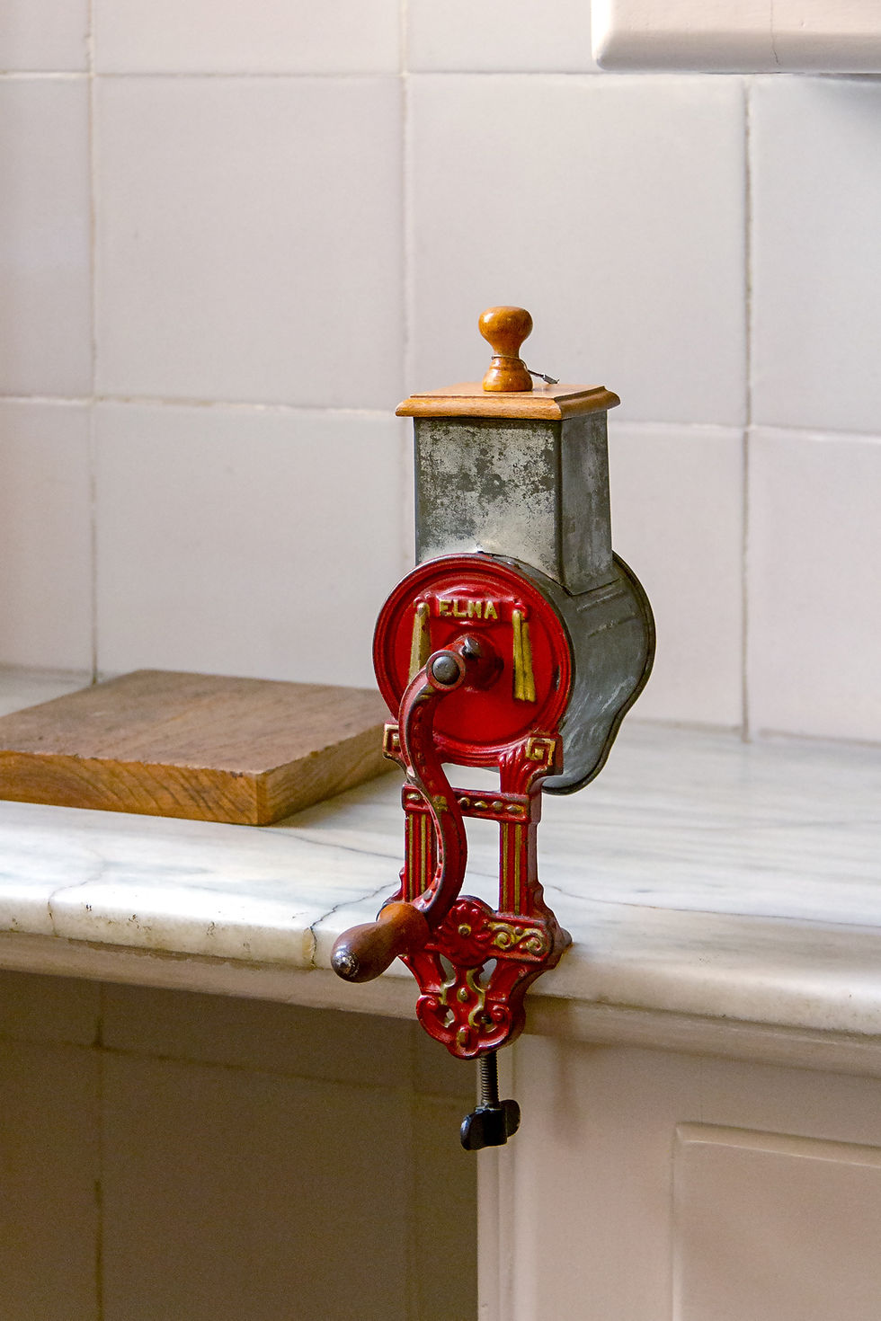 A retro manual coffee grinder, adorned with intricate red and wooden details, is set up on a kitchen counter.