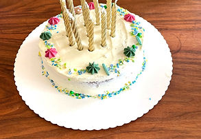 A round white frosted cake decorated with colorful sprinkles, pink and green star-shaped candies, and eight gold spiral candl