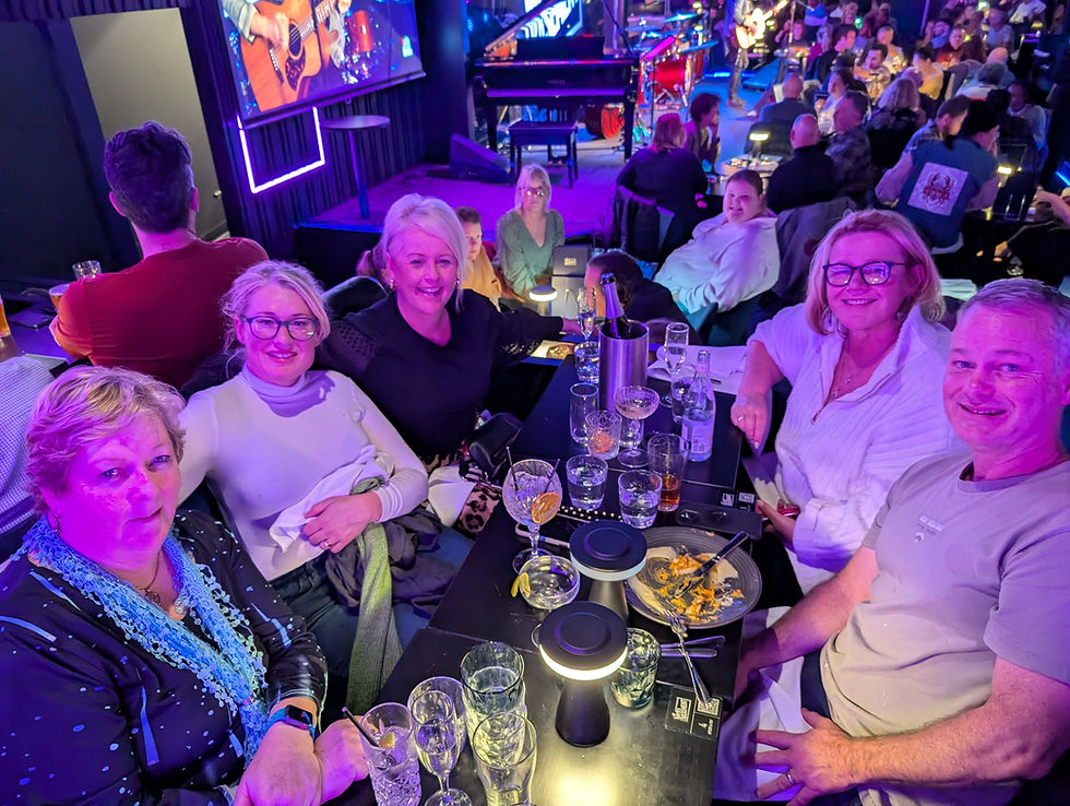 Group of people smiling at a table in a vibrant, crowded venue with colorful lights. Plates and drinks on the table, live music in the background.
