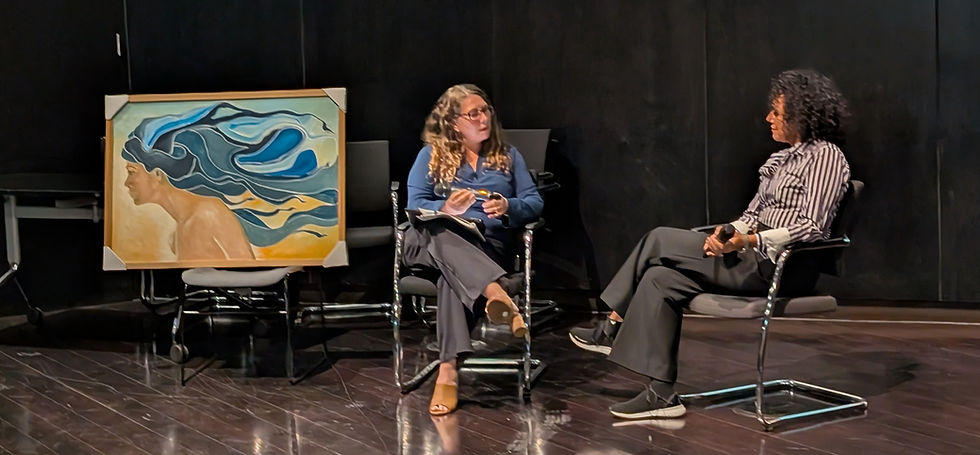 Two people seated on stage discussing art. A vibrant painting with blue, yellow, and orange hues leans against a dark wall.