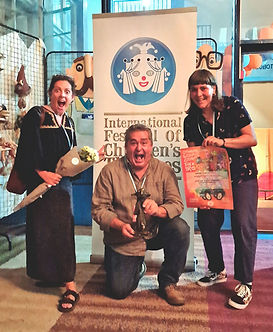 Photo of three people posing joyfully with an award trophy and a certificate in front of a banner for the International Festival of Children's Theaters.