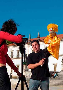 Gil em filmagem no exterior a operar uma marioneta amarela enquanto é filmado por uma operadora de câmara.