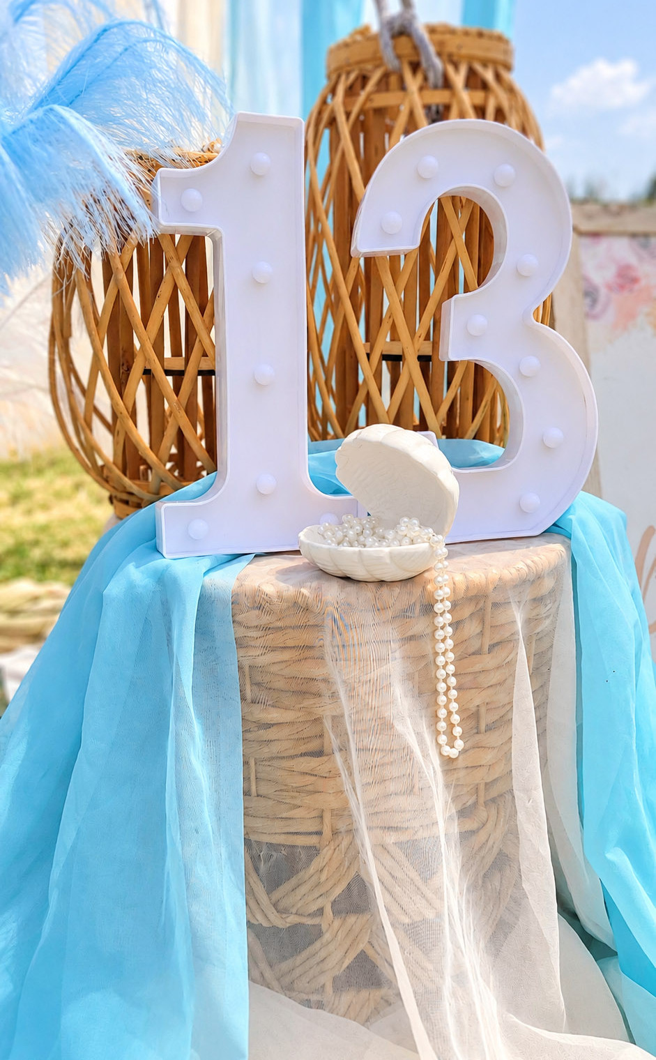 Decorative white number 13 with woven lanterns, blue fabric and seashell pearls at a coastal birthday picnic in San Diego.