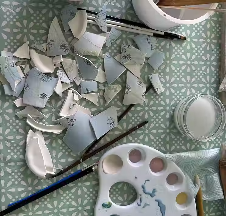 Top-down overhead view of broken ceramic pieces with floral illustrations lying on a green patterned table next to paintbrushes, a liquid jar, and a color palette