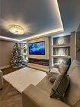 Living room decorated for Christmas with a modern media wall, glowing electric fireplace, white rug, and decorated Christmas tree.