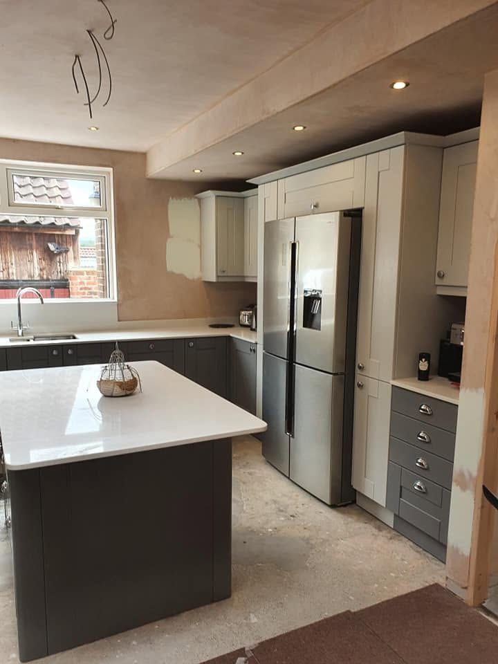 Two-tone shaker kitchen installation featuring grey and off-white cabinets, quartz-topped island, stainless steel American fridge, and unpainted plastered walls.