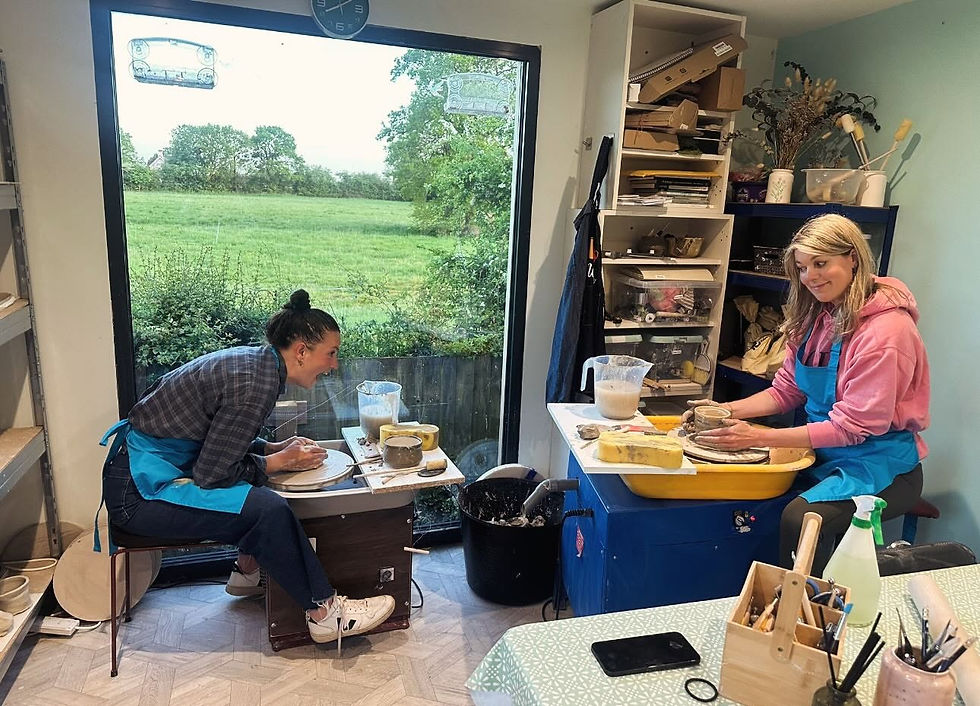 Two women smiling while working on pottery wheels inside a bright studio with a large window looking out at green countryside fields