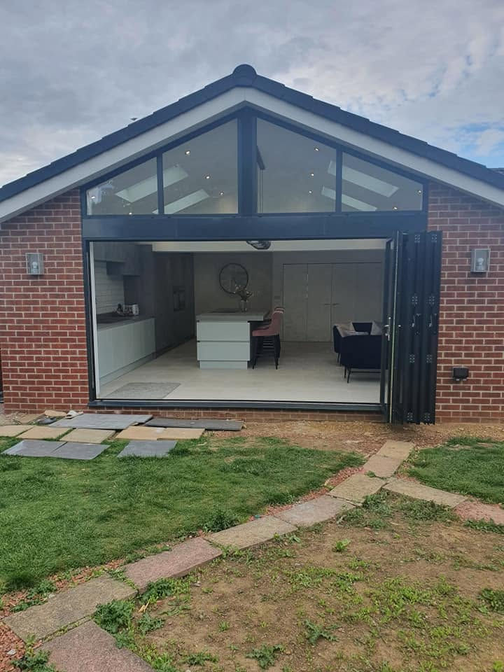 Modern red brick home extension featuring full-width black bi-fold doors, vaulted apex glazed window, and open-plan kitchen interior.