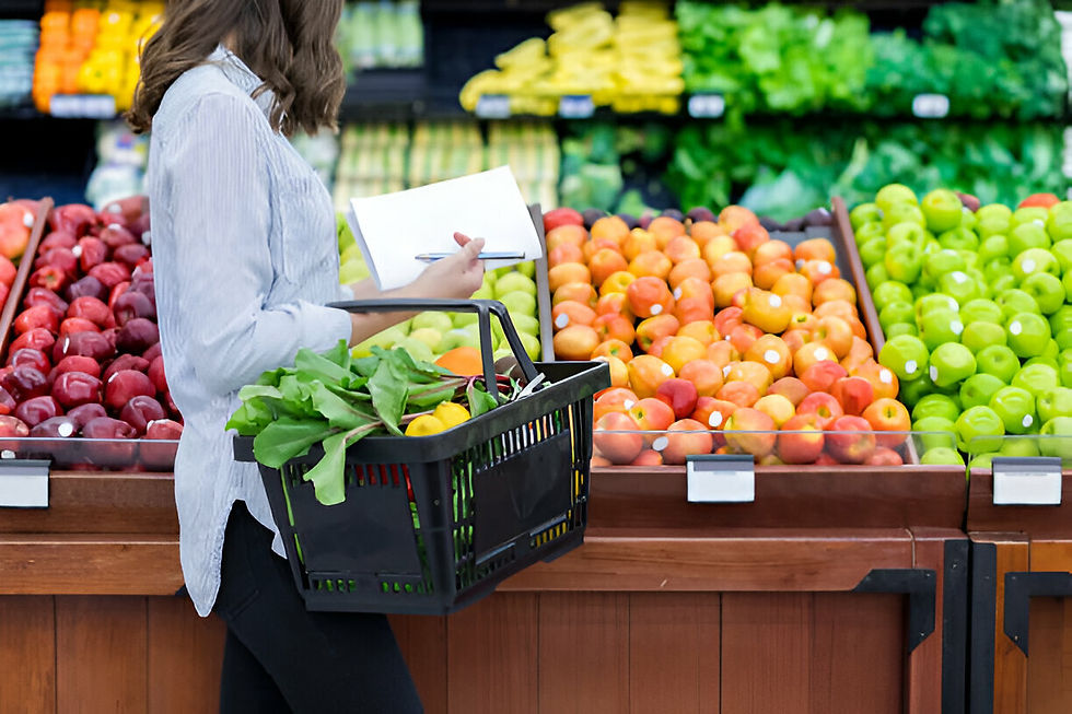 Woman shopping in produce aisle, holding a basket of greens and a notepad. Colorful fruits and vegetables displayed in the background.