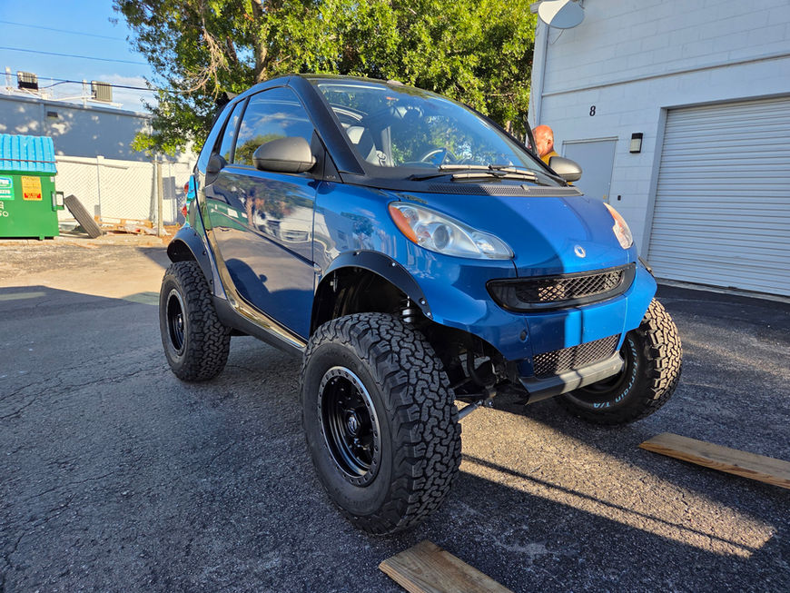 Blue lifted Smart Car with large tires parked in front of a building.