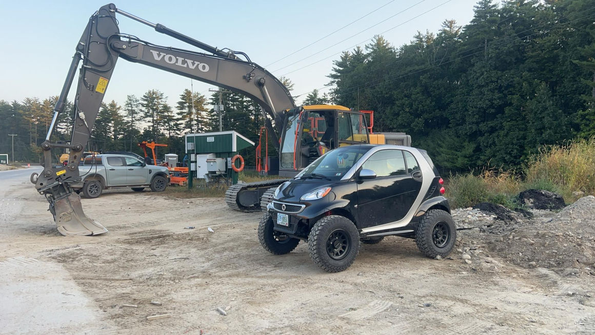 Black Lifted Smart car, Volvo excavator, and other construction equipment on site. BRAAP Smart Car Lift Kits