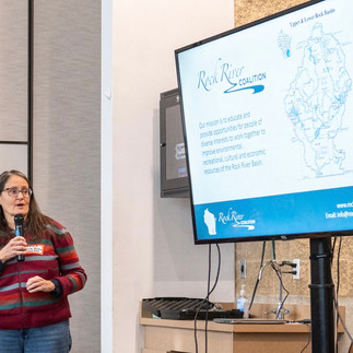 Becca Dymzarov holds a microphone and looks toward a large screen showing information on the Rock River Coalition