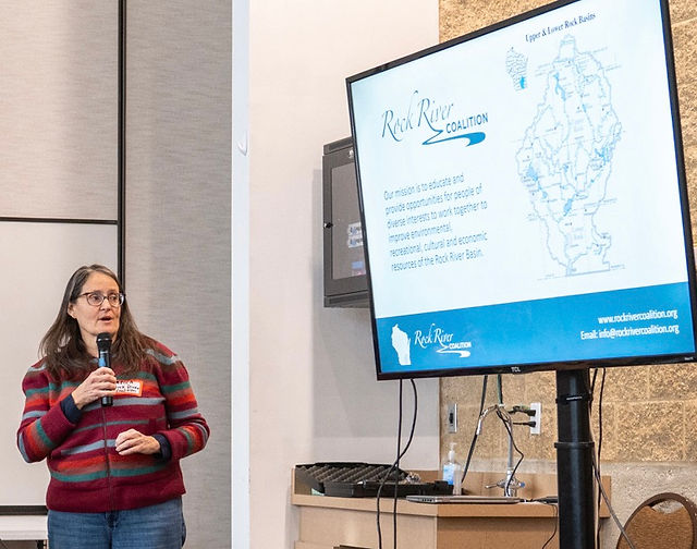 Becca Dymzarov holds a microphone and looks toward a large screen showing information on the Rock River Coalition 