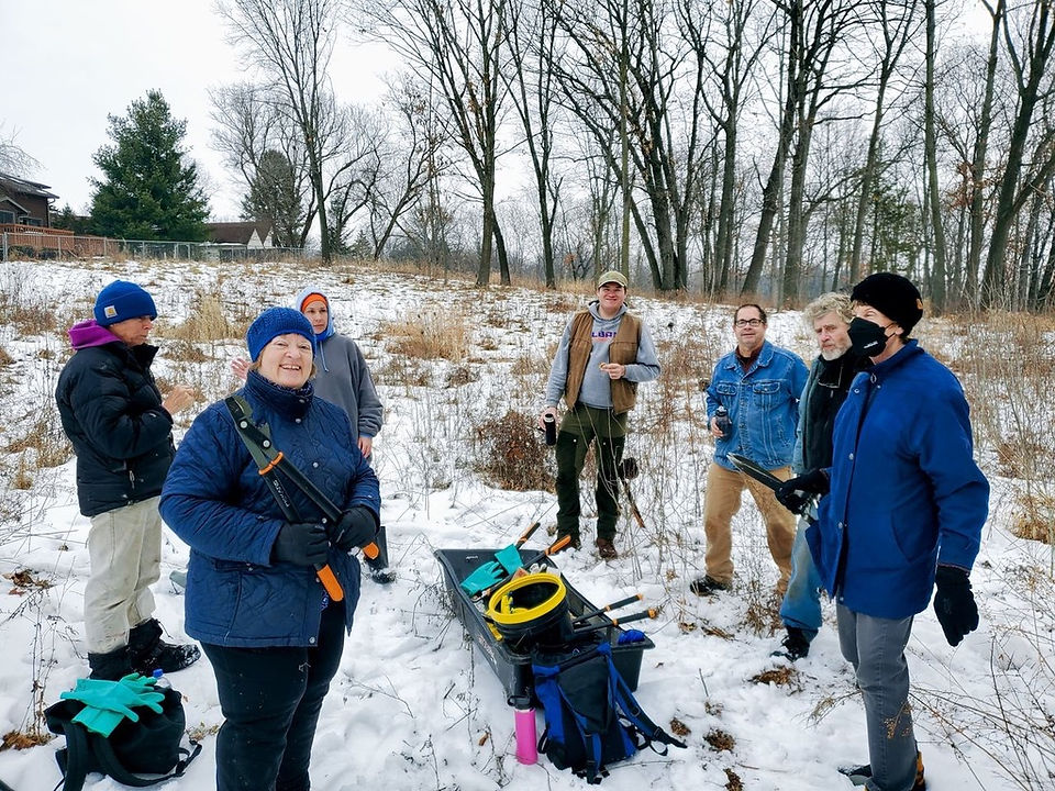 Seven warmly dressed volunteers (men and women) stand in a snowy prairie with woods behind them. They have a sled full of tools for cutting brush.