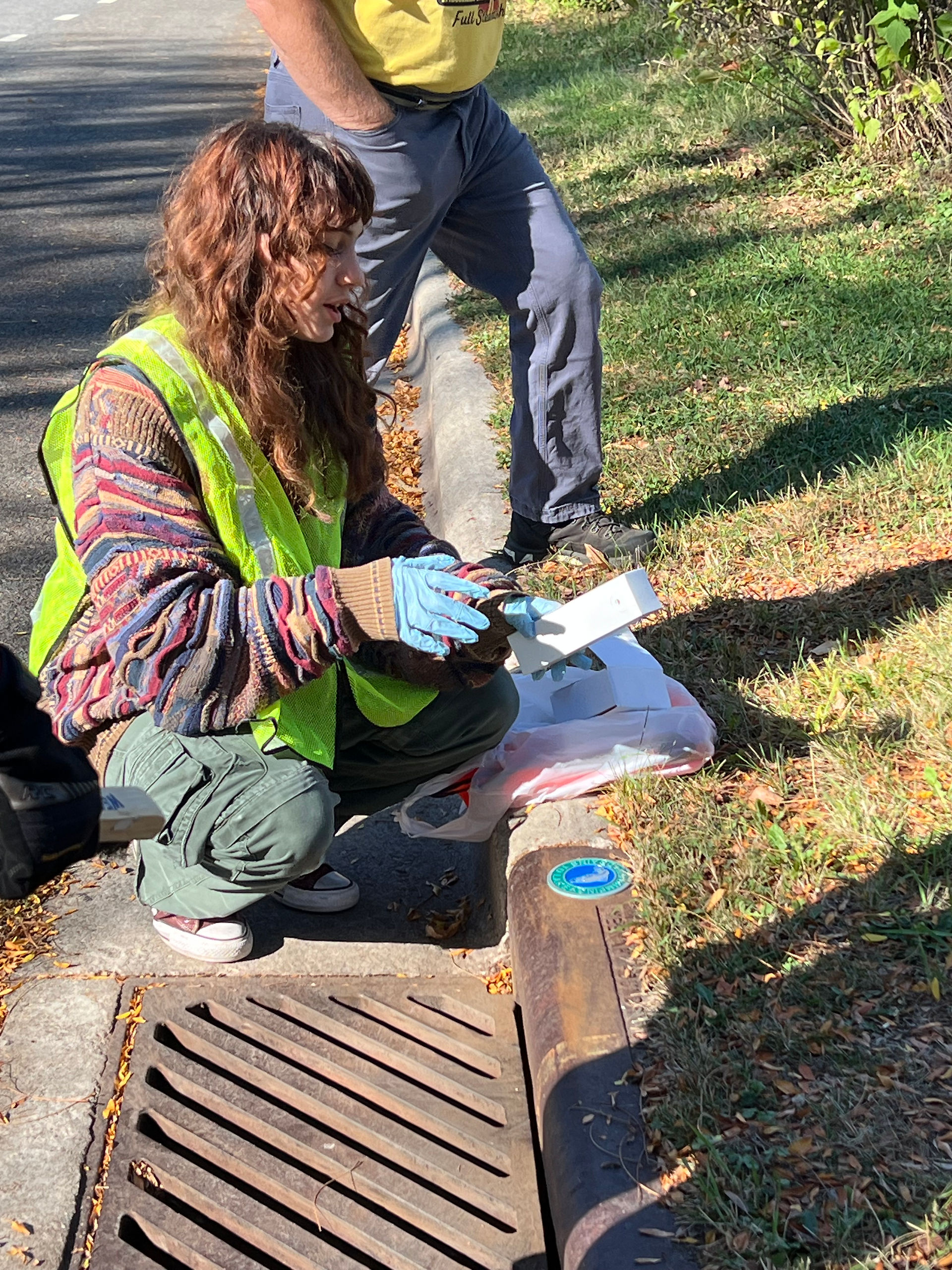 A teenage girl with long curly brown hair kneels by a stormwater drain, wearing a high-visibility yellow safety vest and demonstrating how to install a storm drain marker.