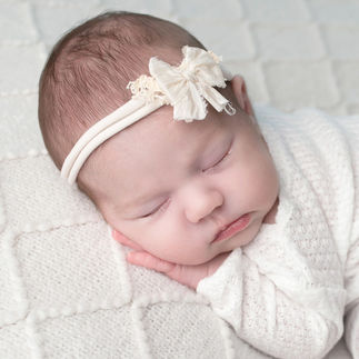 Close-up of a newborn baby girl's face, wearing a white flower headband, with eyes closed, resting on a white knit blanket. Taken by Sacramento maternity and newborn photographer Kristina Martin