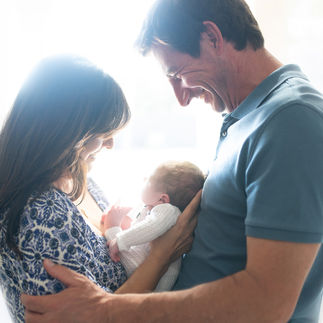 Parents standing by a window, looking down lovingly at their newborn baby girl, with sunlight streaming in behind them. Beautifully backlit family picture. Taken by Sacramento maternity and newborn photographer Kristina Martin