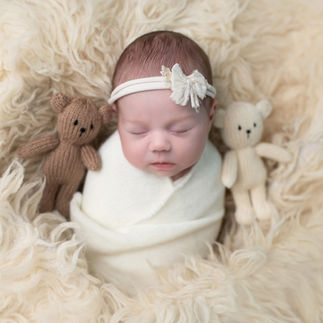 Newborn baby girl wrapped in a white swaddle, holding two small teddy bears, lying on a cream-colored furry blanket. Taken by Sacramento maternity and newborn photographer Kristina Martin
