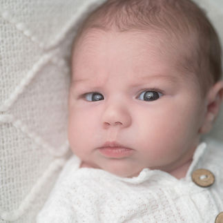 Newborn baby girl with a serious expression, wearing a white knitted outfit and flower headband. Taken by Sacramento maternity and newborn photographer Kristina Martin