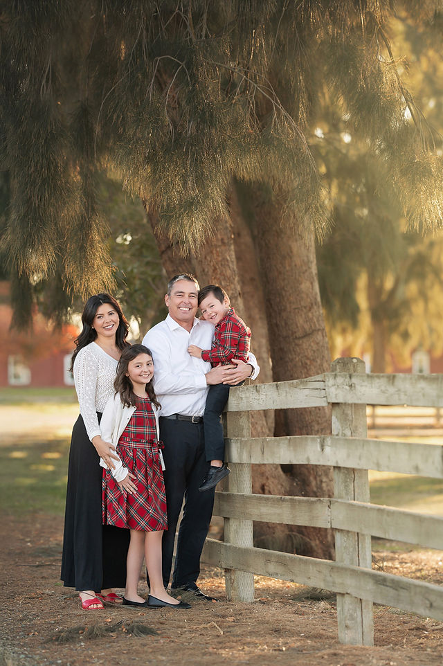 A family session photographed by a Sacramento family photographer at an outdoor location featuring a wooden fence and warm, golden light. The family includes a father, mother, and two children dressed in coordinating red and white outfits. The photos capture a mix of candid and posed moments, highlighting their connections and playful interactions, set against a serene, tree-lined backdrop.