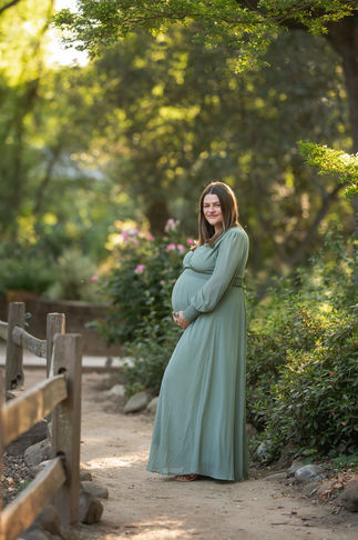 Pregnant woman in a green dress standing near a wooden fence, cradling her belly in a serene outdoor setting taken by Sacramento photographer Kristina Martin.