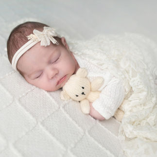 Newborn baby girl wrapped in a white swaddle, holding a small teddy bear, resting on a cream-colored furry blanket. Taken by Sacramento maternity and newborn photographer Kristina Martin