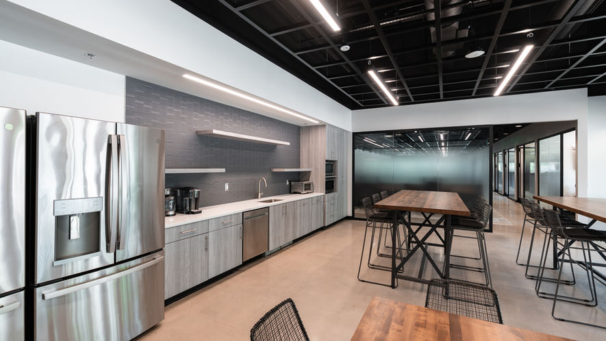 Modern office kitchen with stainless steel appliances and wooden tables and chairs.