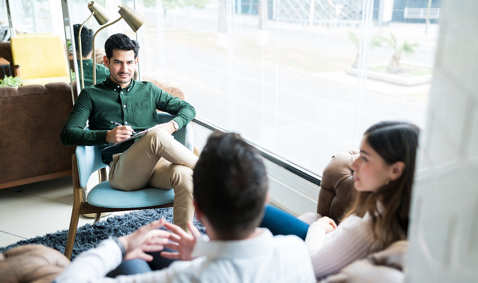 Therapist in green shirt holds pen and clipboard, listening to couple on couch in modern, bright office. Mood is professional and calm.