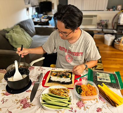 A man in a gray shirt sits at a table and assembles his kimbap. The kimbap ingredients are all over the table.