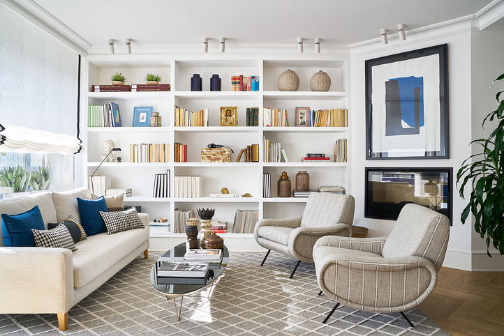 Fotografía de interiorismo de este salón con muchísima luz natural. Se ve la chimenea a un lado y la gran librería de obra que preside el espacio. 