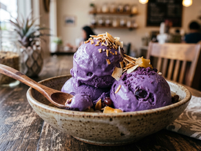 Bowl of purple ube ice cream topped with coconut flakes and nuts on a wooden table in a cozy café, with blurred diners behind.