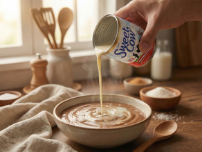 A hand pouring Sweet Cow evaporated milk from a can into a steaming bowl of savory soup in a sunlit kitchen.