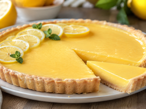 A beautiful, golden-yellow lemon tart with a fluted crust, decorated with fresh lemon slices and mint leaves, with one slice neatly cut out on a wooden table.