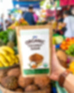 A hand holding a bag of Jans Organic Coconut Sugar in a vibrant outdoor market with coconuts and other fruits in the background.