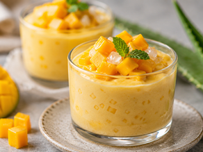 Two elegant glass cups filled with a pale yellow Jans Aloe Fruit Mousse, topped with vibrant diced fresh mango chunks, small translucent aloe vera cubes, and a fresh mint sprig, next to a sliced mango and an aloe leaf.