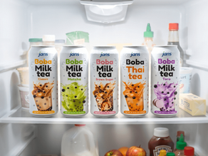 Five colorful cans of Jans Boba Milk Tea—Classic, Matcha, Brown Sugar, Thai Tea, and Taro chilling on a shelf inside a well-stocked refrigerator.