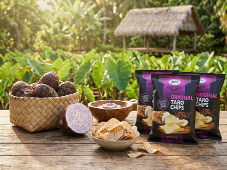 A woven basket filled with raw taro corms sits on a wooden table next to a sliced purple-speckled taro root, a small wooden bowl of dip, and bags of Jans Original Taro Chips, set against a bright, sunlit outdoor taro field and a thatched hut.