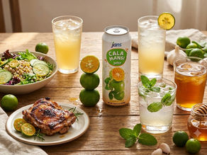 A table spread showcasing different ways to use calamansi juice, featuring a can of Jans Cala Mansi Juice, tall glasses of iced cocktails, a fresh green salad, and a plate of grilled chicken.
