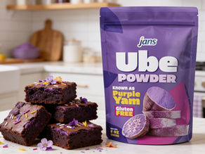 A stack of dense, fudgy purple ube brownies decorated with purple sprinkles and edible flowers, displayed on a kitchen counter next to a purple stand-up pouch of Jans Ube Powder.