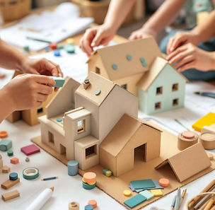 Children collaboratively building cardboard homes and neighborhood structures using loose parts, construction materials, and simple design tools during a cozy preschool engineering and creative building activity with warm natural light