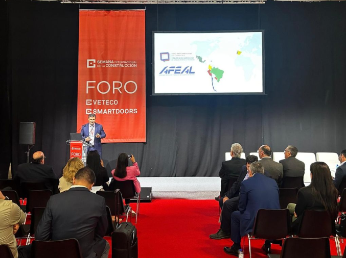 Palestra com palco preto e faixa laranja com nome do evento, palestrante de terno e público sentado de costas