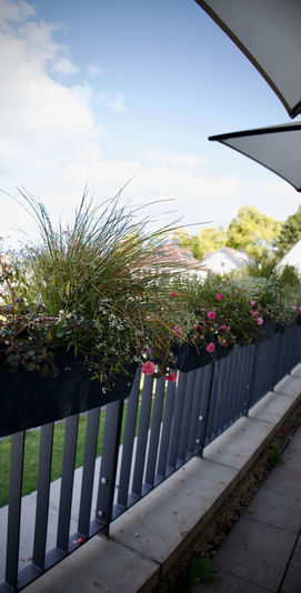 Moderner Balkon mit Blumenkästen, grünen Pflanzen und rosa Blüten unter blauem Himmel.